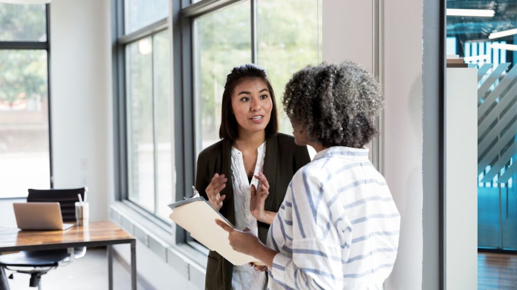 Two colleagues in conversation beside a window in an office, one holding a clipboard