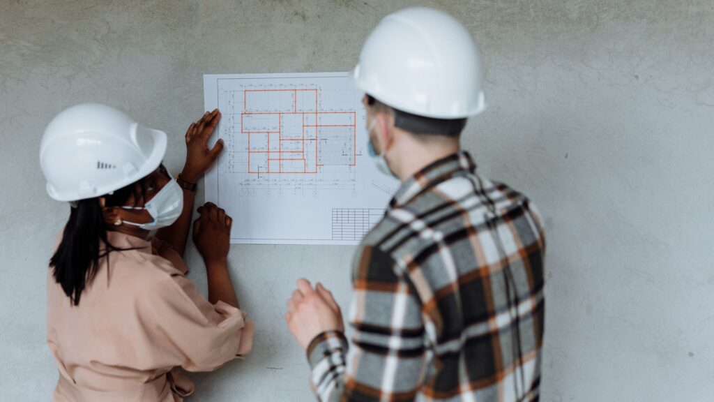 Two people in hard hats holding rolled drawings inside a timber-framed building under construction