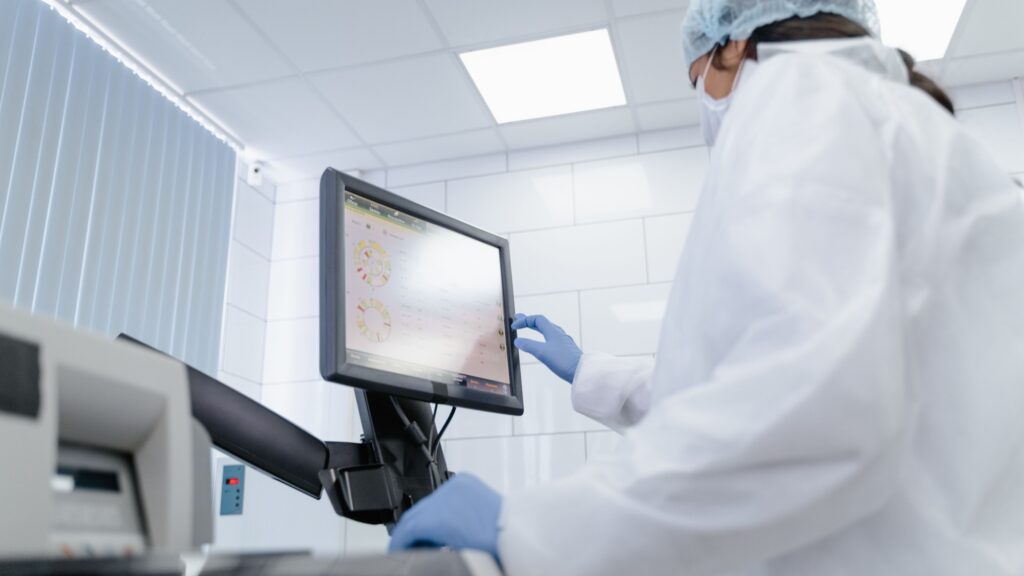 A gowned worker in a hairnet and gloves operating a touchscreen terminal in a production cleanroom