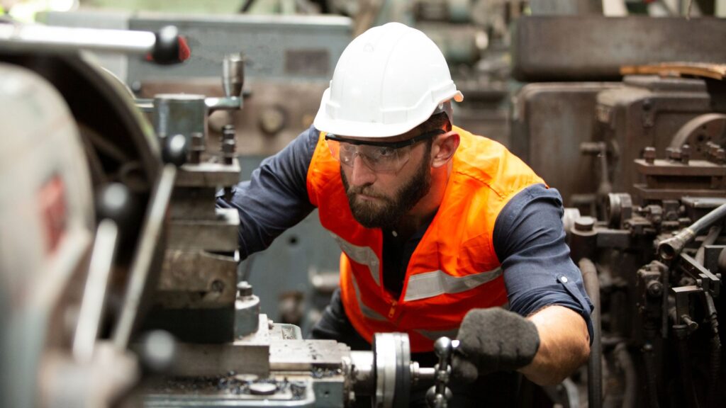 A machinist in safety glasses and an orange high-visibility vest leaning in to work at a lathe