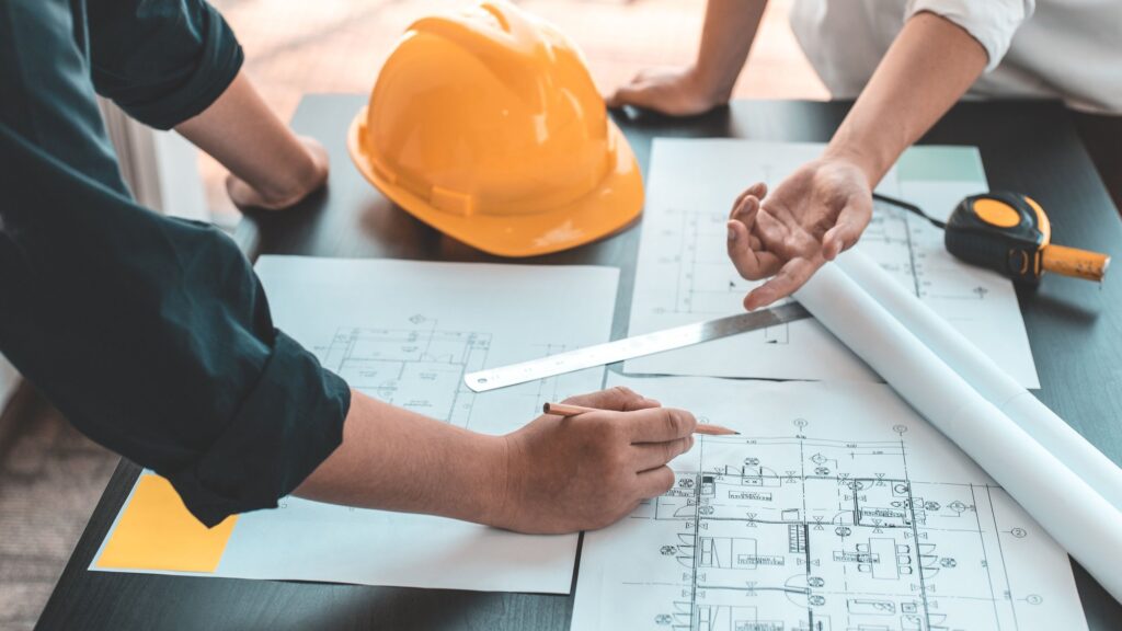 Two people's hands over construction drawings on a table, beside an orange hard hat, a ruler and a tape measure