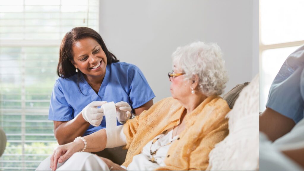 A nurse in blue scrubs and gloves bandaging the arm of an older woman sitting beside her