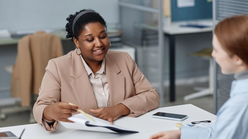A woman in a blazer reading a document across a desk from a colleague in an office