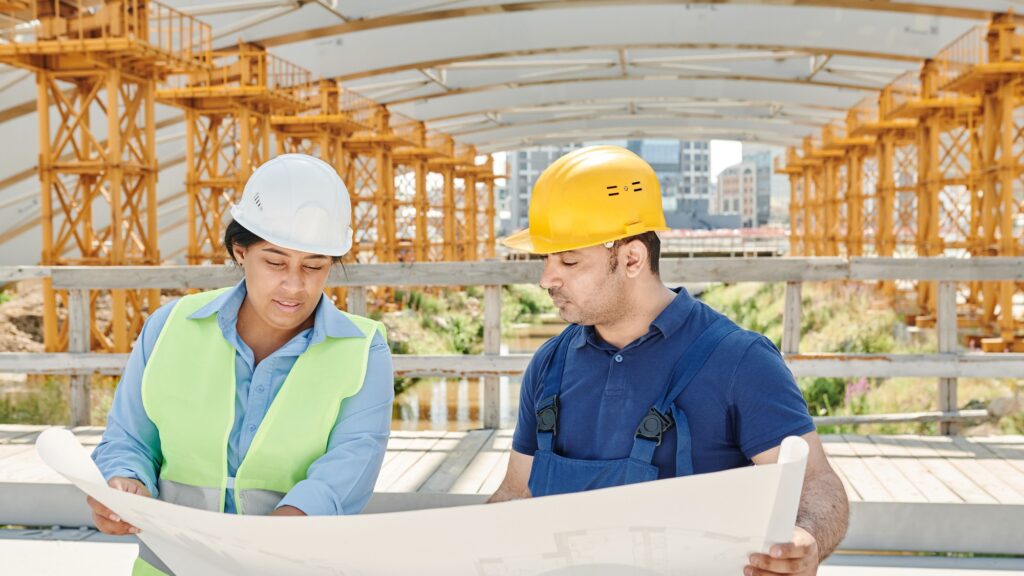 Two workers in hard hats studying a large drawing under a yellow steel falsework frame