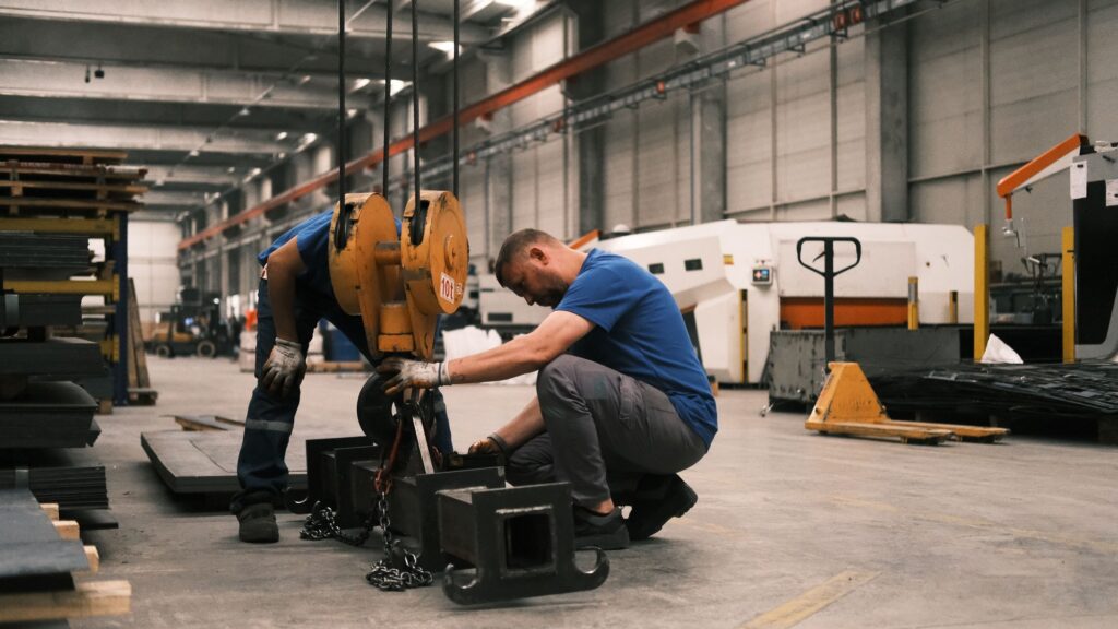 Two workers kneeling to adjust heavy lifting equipment on a manufacturing floor