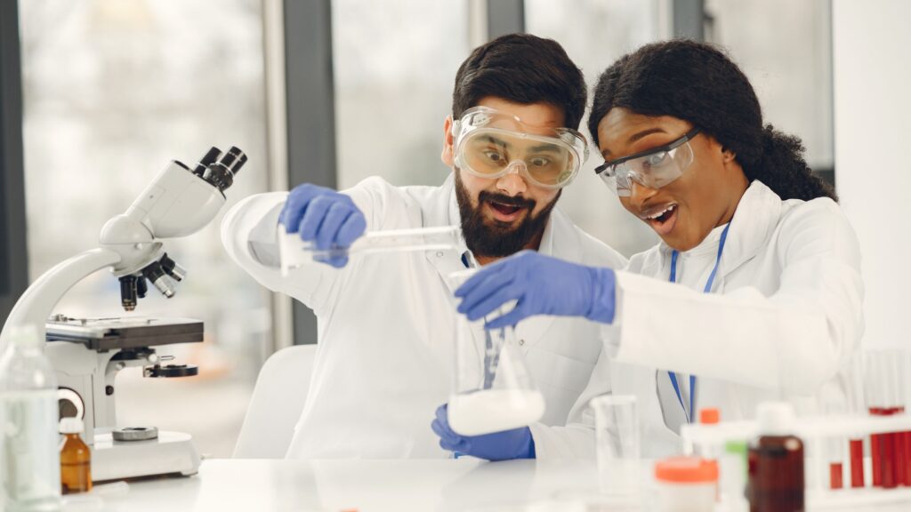 Two researchers in laboratory coats, goggles and gloves working with glassware at a bench
