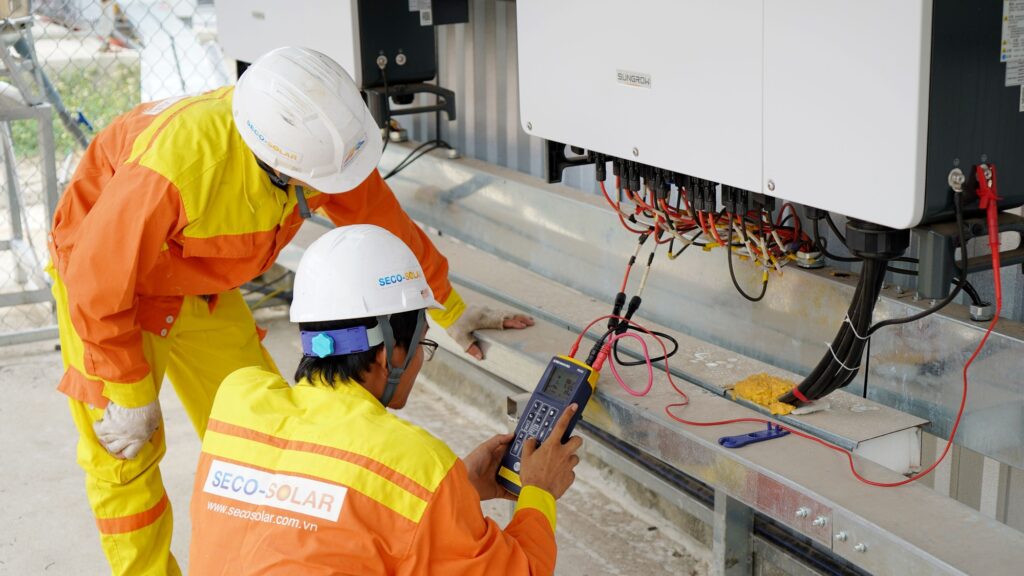Two workers in orange high-visibility overalls testing a wiring cabinet with a handheld meter