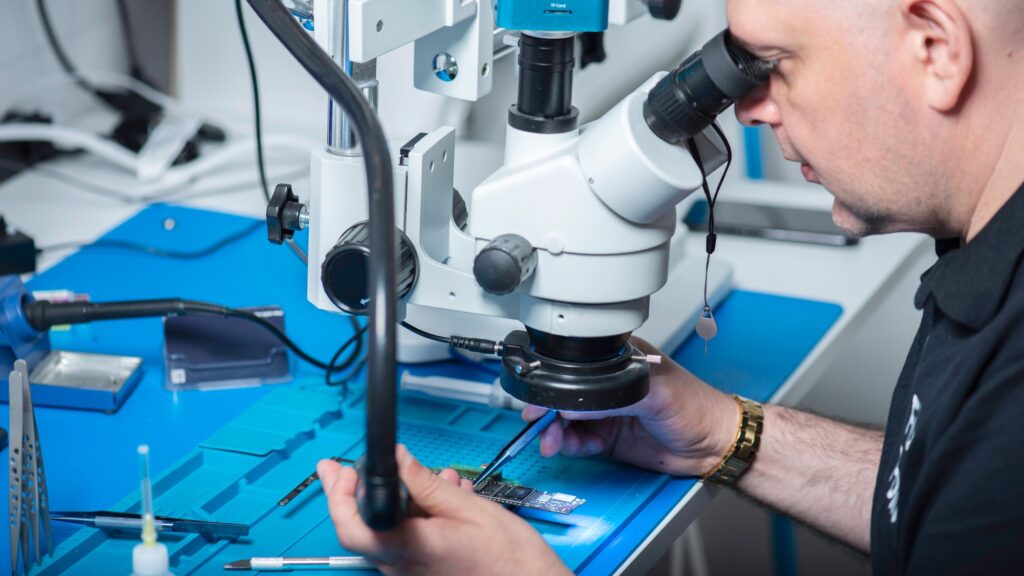 A stereo microscope over a circuit board on a blue bench mat, a hand steadying the board beneath it
