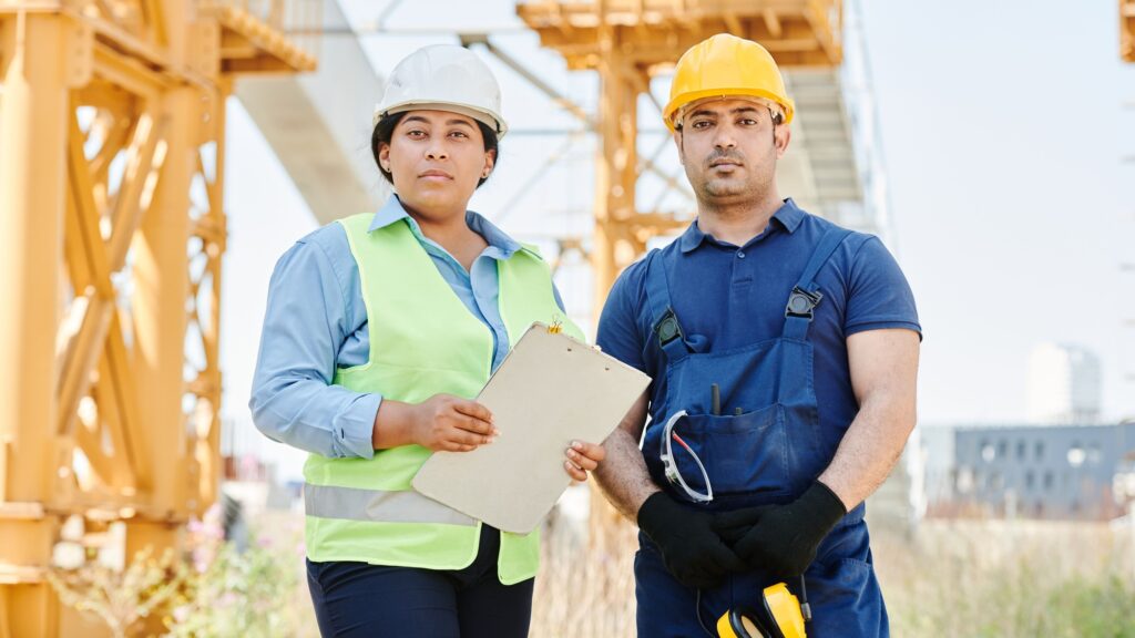 Two engineers in hard hats and high-visibility clothing talking on an industrial site, a crane behind them
