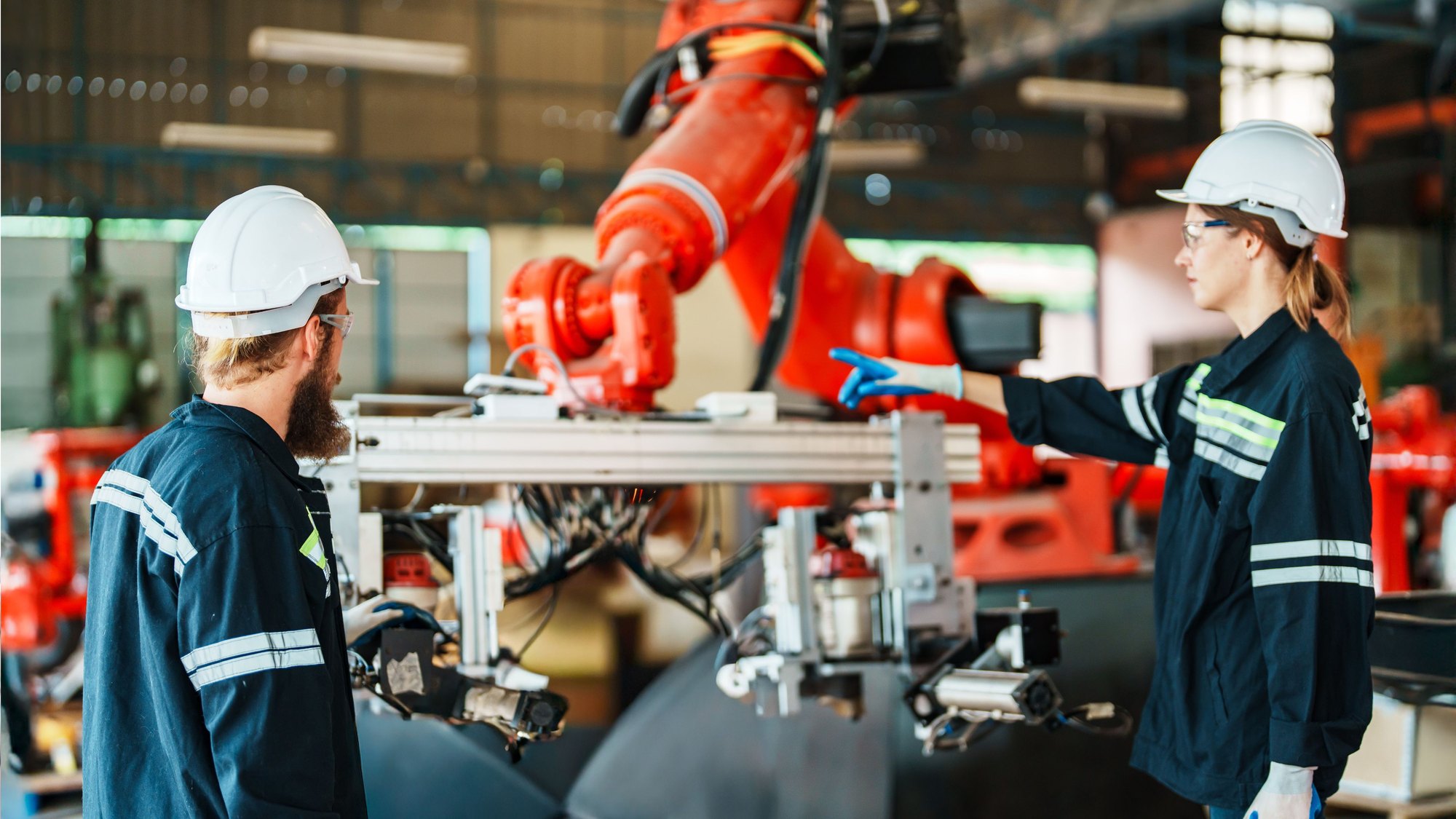 Two workers in hard hats and dark coveralls operating a large red industrial robot on a factory floor