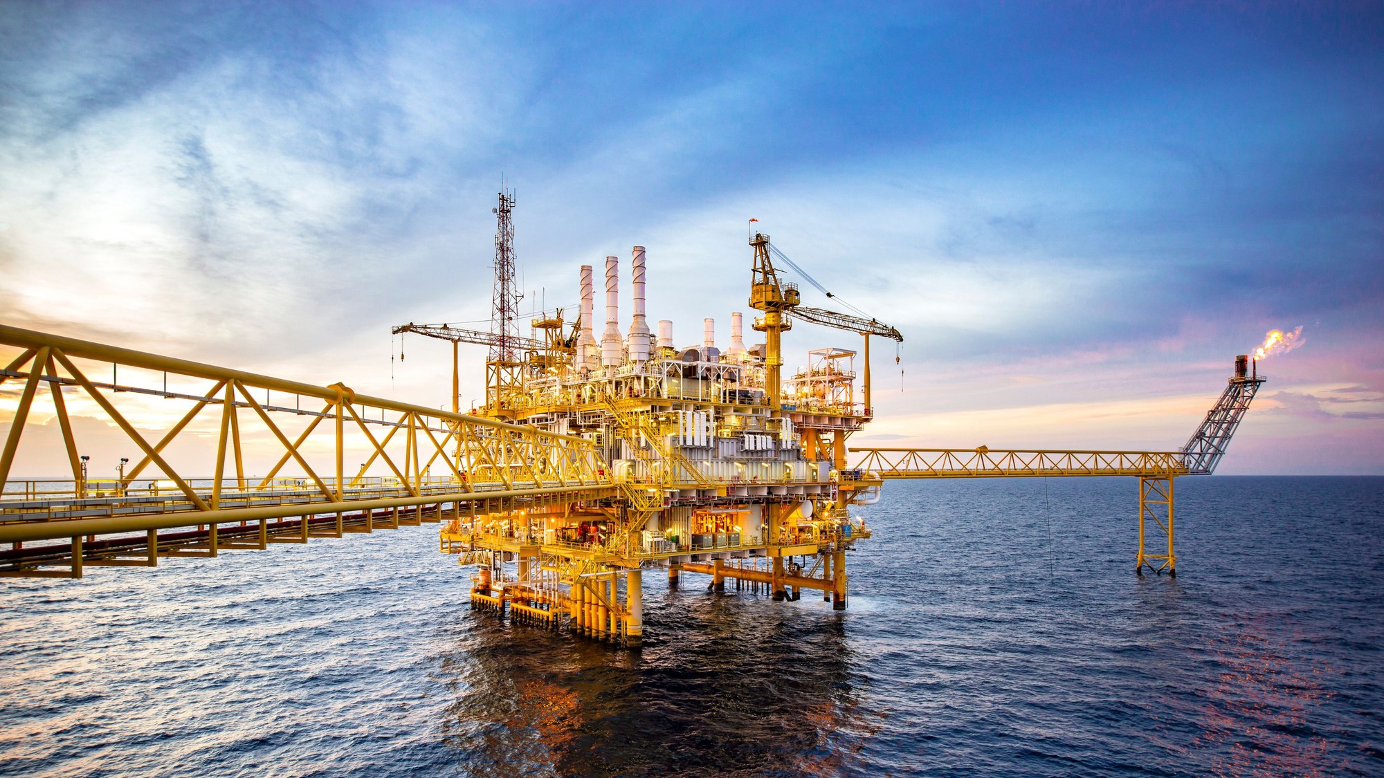 An offshore oil and gas platform at sea at dusk, its flare burning at the end of a long bridge