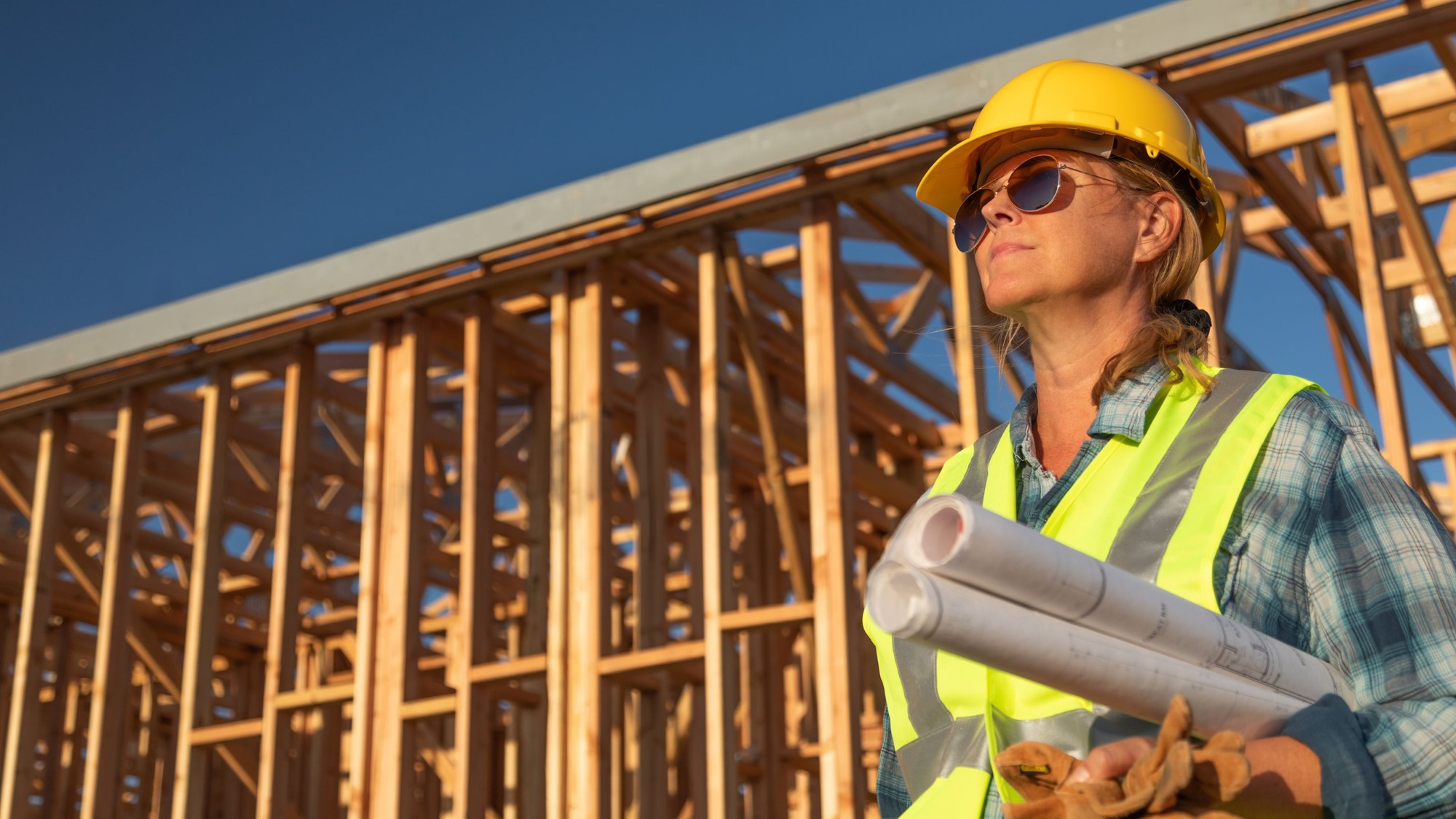 A worker in a yellow hard hat and high-visibility vest carrying rolled drawings inside a timber-framed building under construction