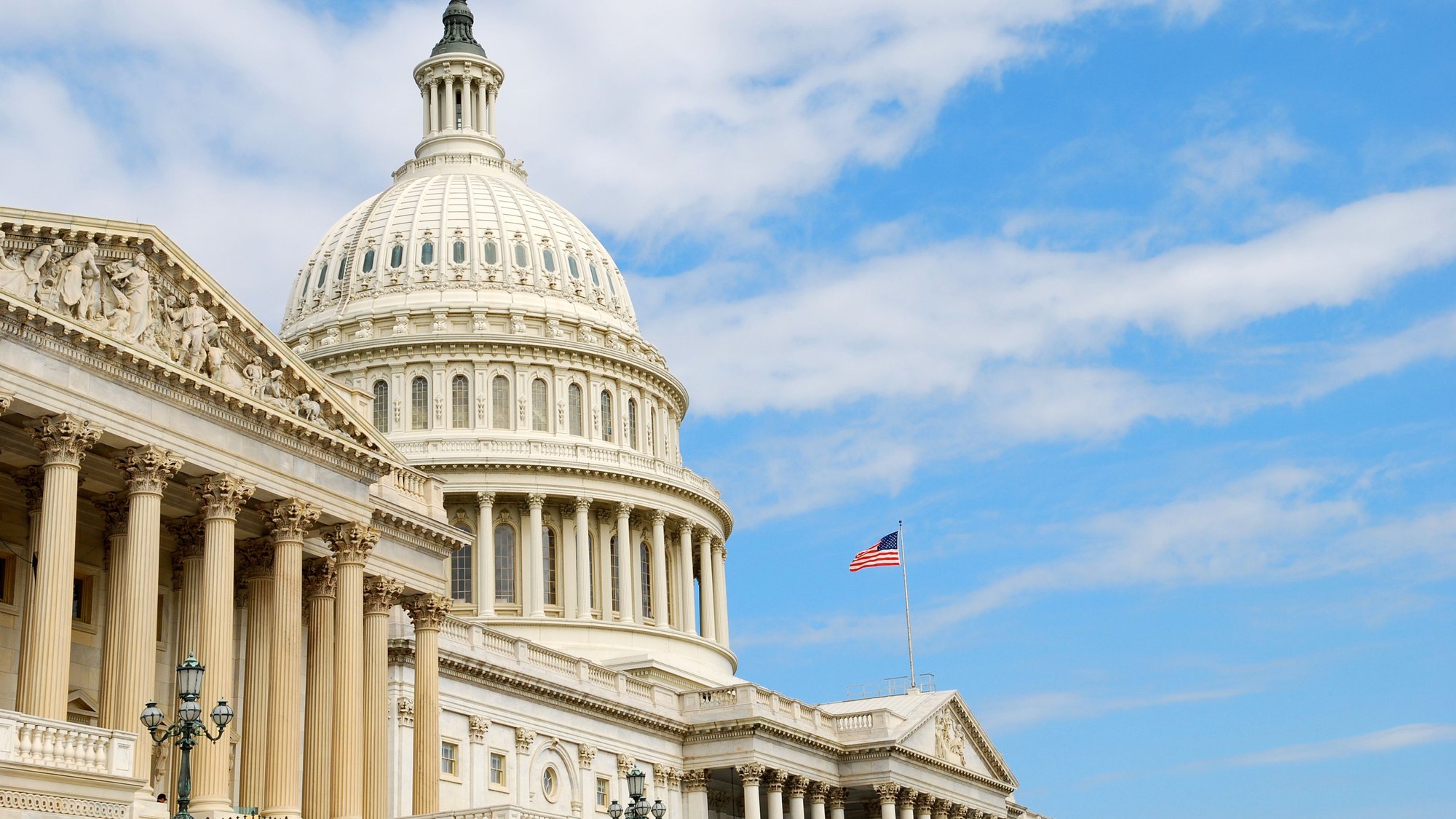 The dome of the United States Capitol against a blue sky, the flag flying beside it