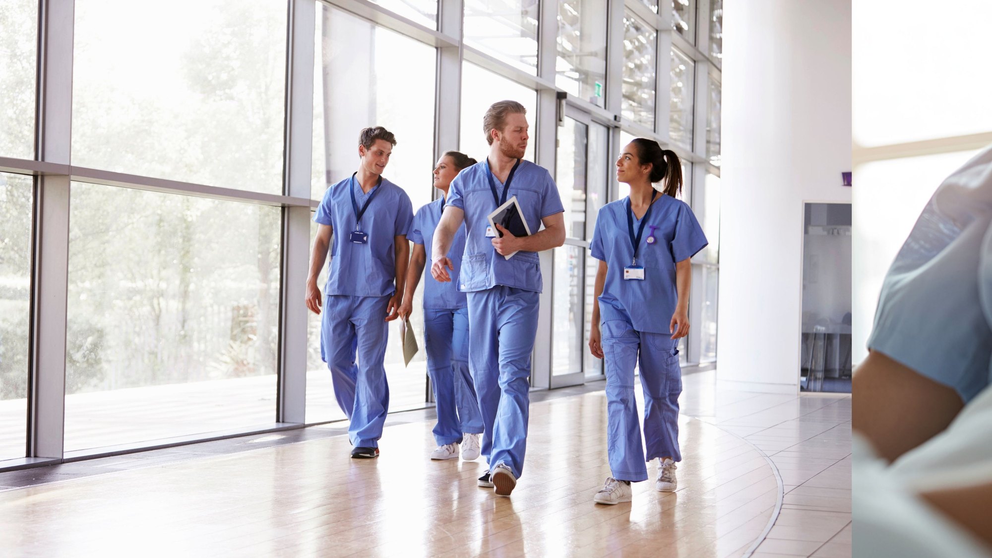 Four clinicians in blue scrubs walking together down a bright, glass-walled hospital corridor