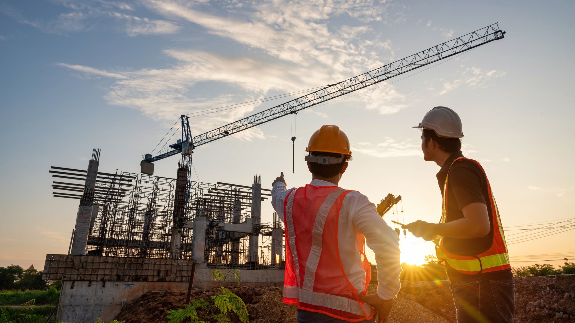 Two workers in high-visibility vests and hard hats on a construction site at sunset, a tower crane behind them