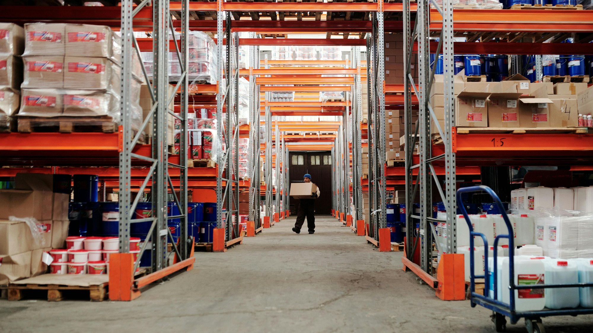 A long warehouse aisle of racked pallets, a worker at the far end
