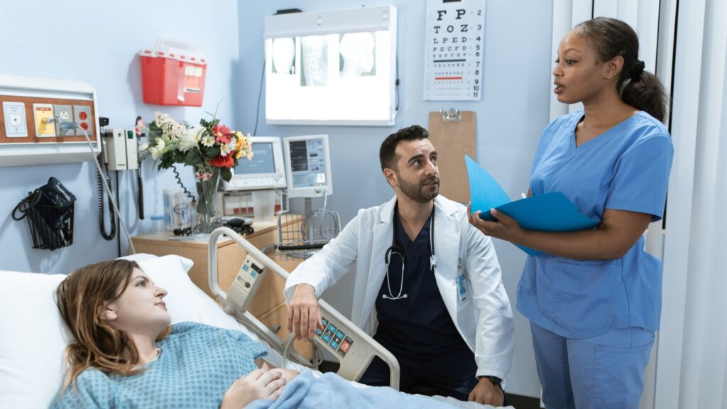 A doctor and two colleagues in scrubs talking at the bedside of a patient on a ward