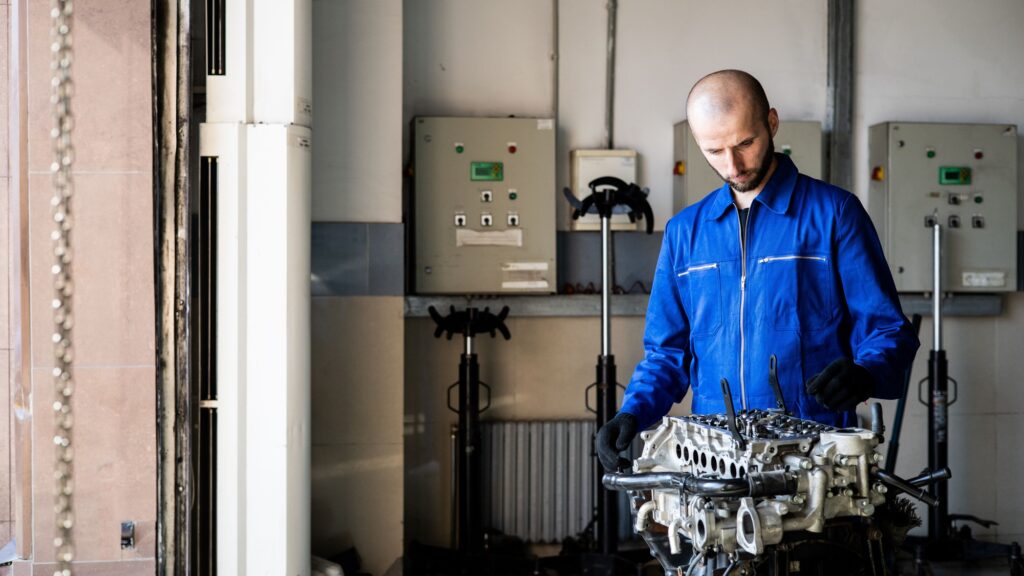 A technician in blue overalls working on an engine block mounted on a workshop stand