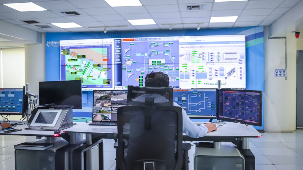 An operator at a control room desk facing a wall of plant mimic displays