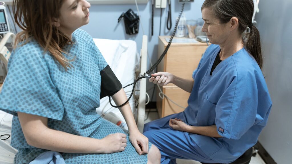A nurse in blue scrubs taking the blood pressure of a seated patient in a gown