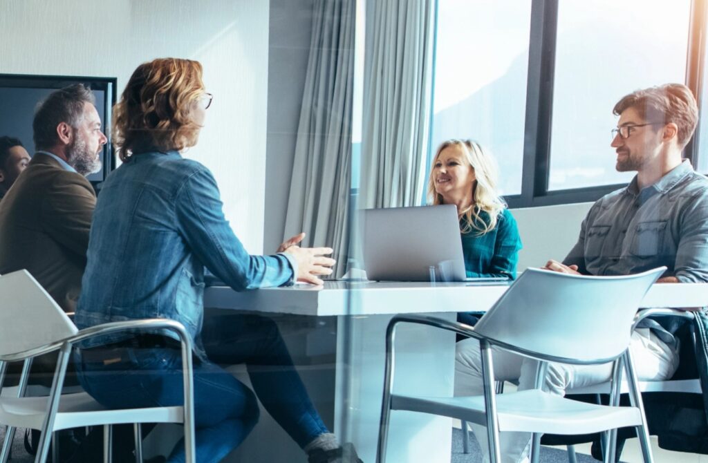 Five colleagues talking around a table in a glass-walled meeting room, a laptop open between them