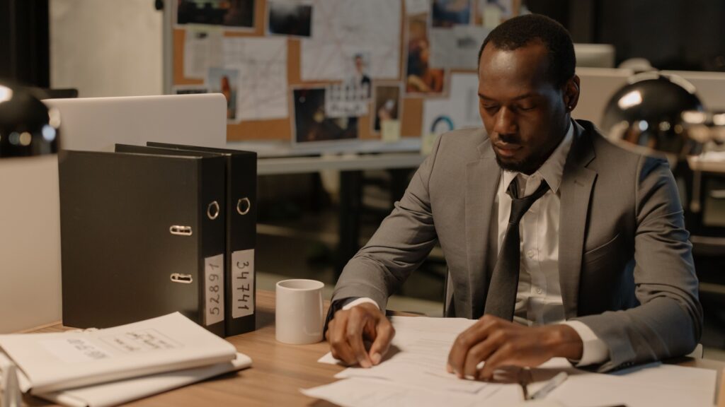 A man in a suit working through paperwork at an office desk beside a row of ring binders