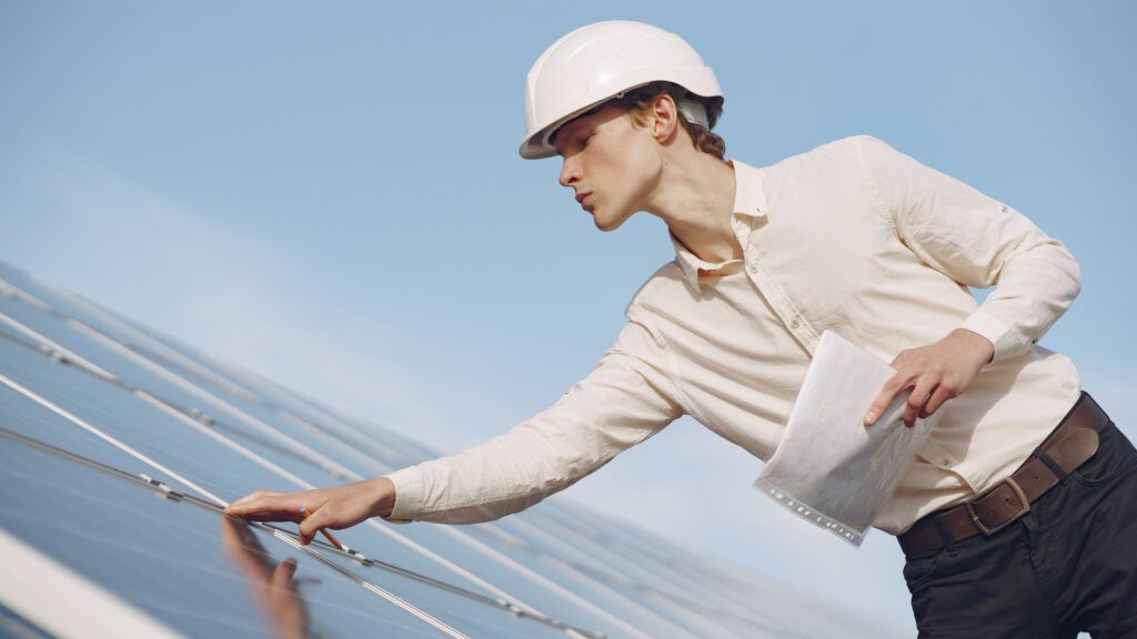 An inspector in a hard hat reaching across a row of solar panels, notes in hand