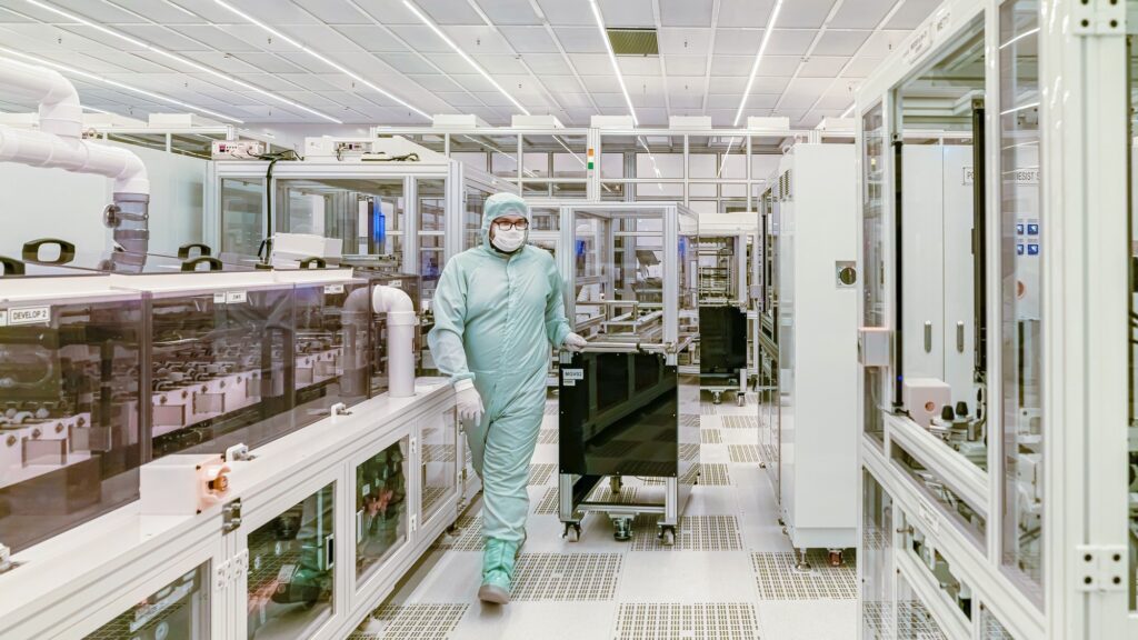 A worker in a full cleanroom suit and mask walking between tool bays in a fabrication plant