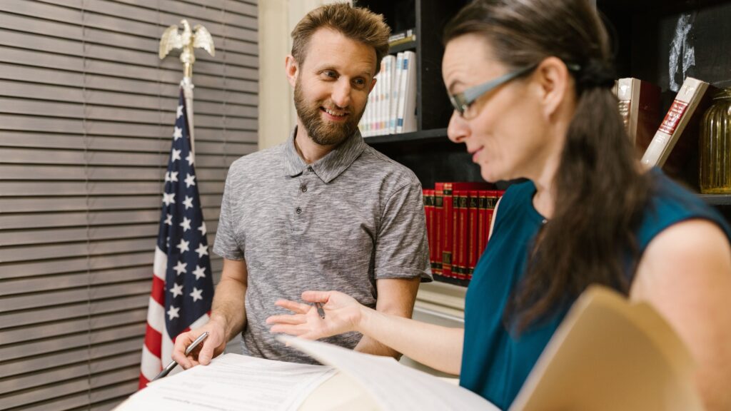 Two colleagues talking over paperwork at a desk, a United States flag on the wall behind them