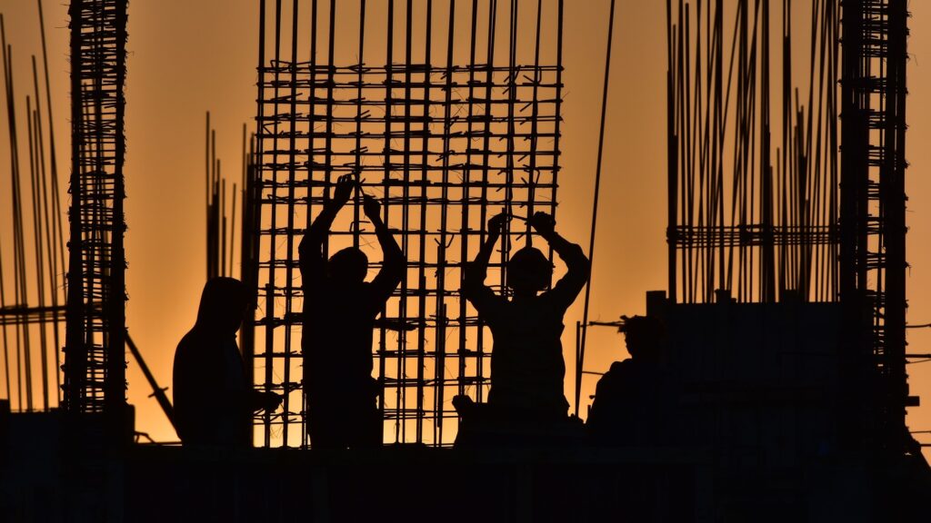 Workers silhouetted against an orange sky as they tie a tall grid of reinforcing bar