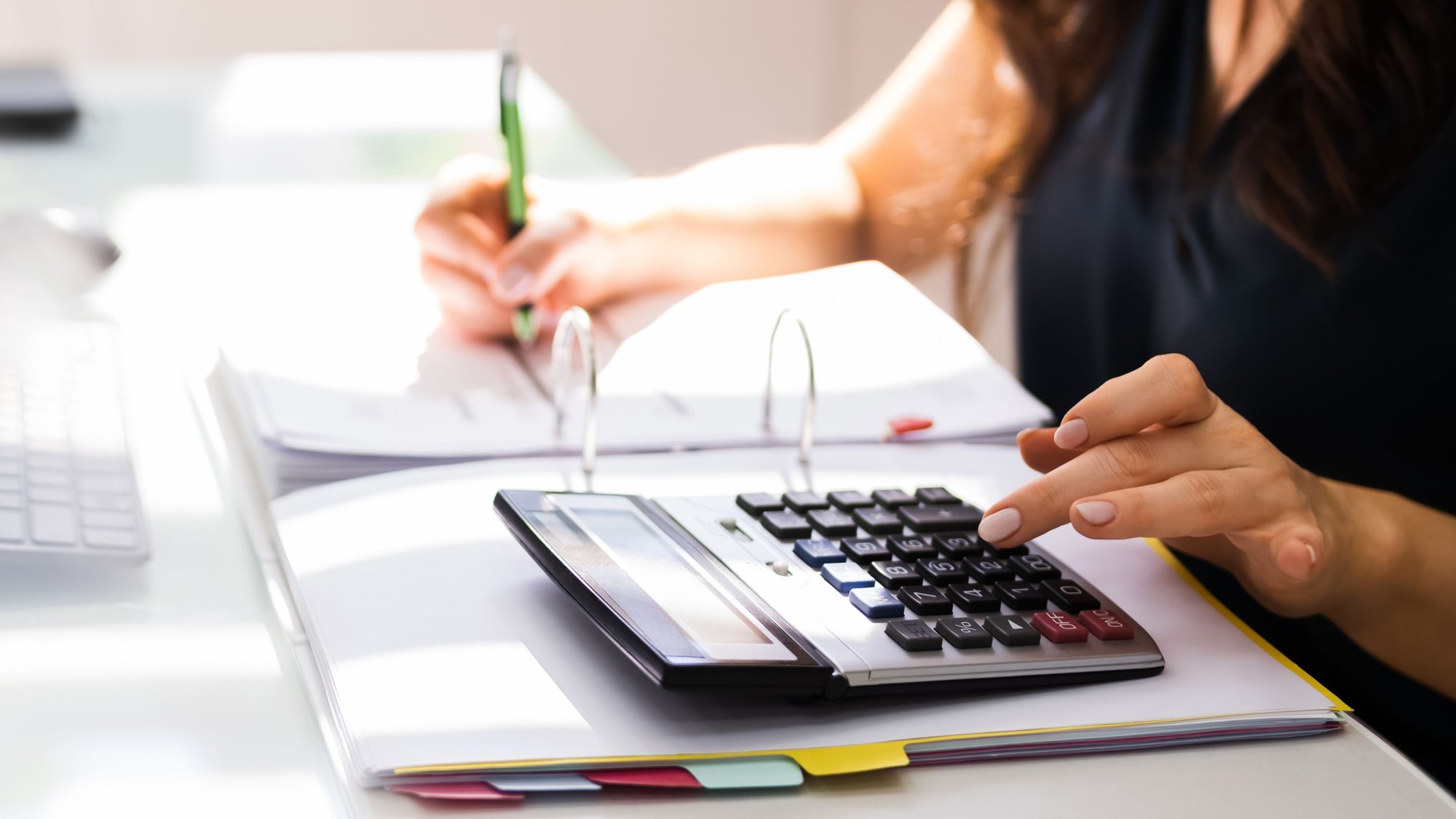 Hands writing in a ledger beside a calculator and a bound report