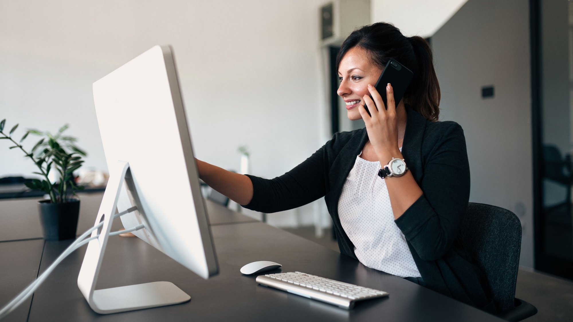 A woman at her desk taking a phone call and reaching for her computer