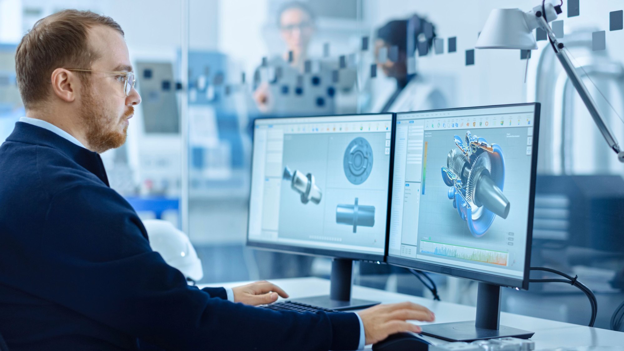 A man working at two monitors showing three-dimensional CAD models of a machined part