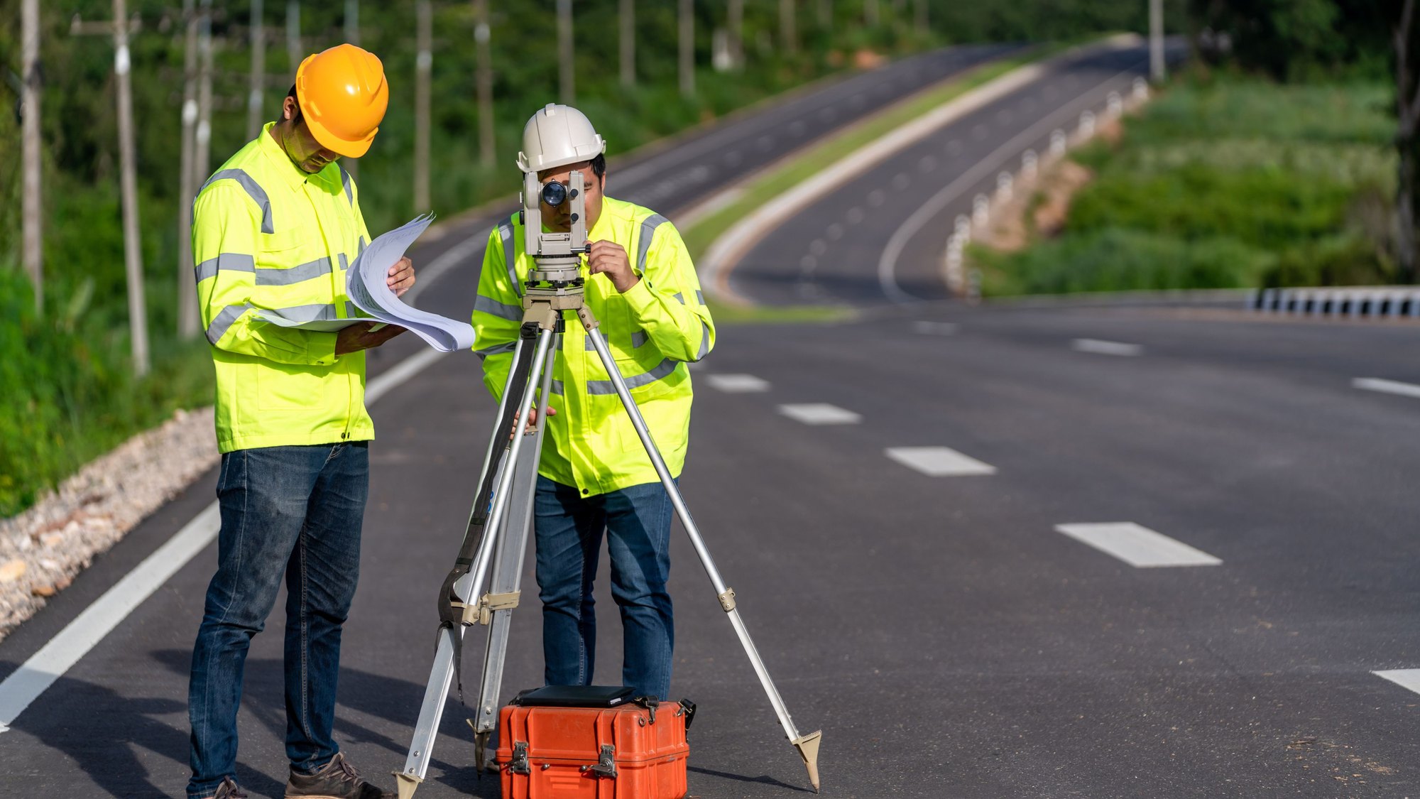 Two surveyors in high-visibility vests and hard hats setting up a total station at the edge of a new road