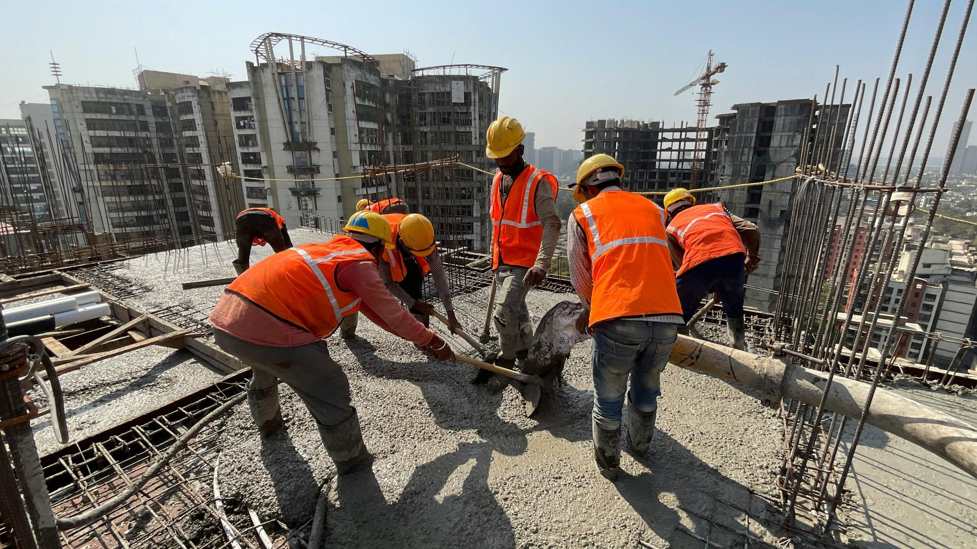 Workers in orange high-visibility vests pouring and levelling concrete on a high-rise deck, a tower crane behind them