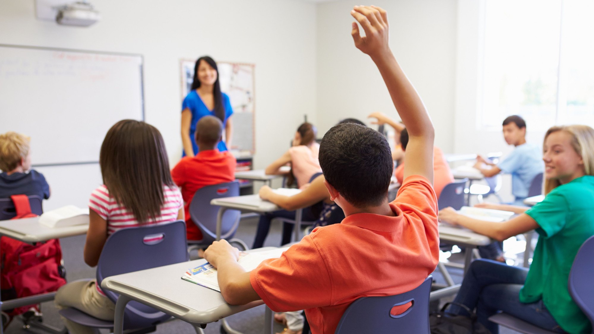 A classroom of children at their desks with one hand raised, the teacher standing at the front