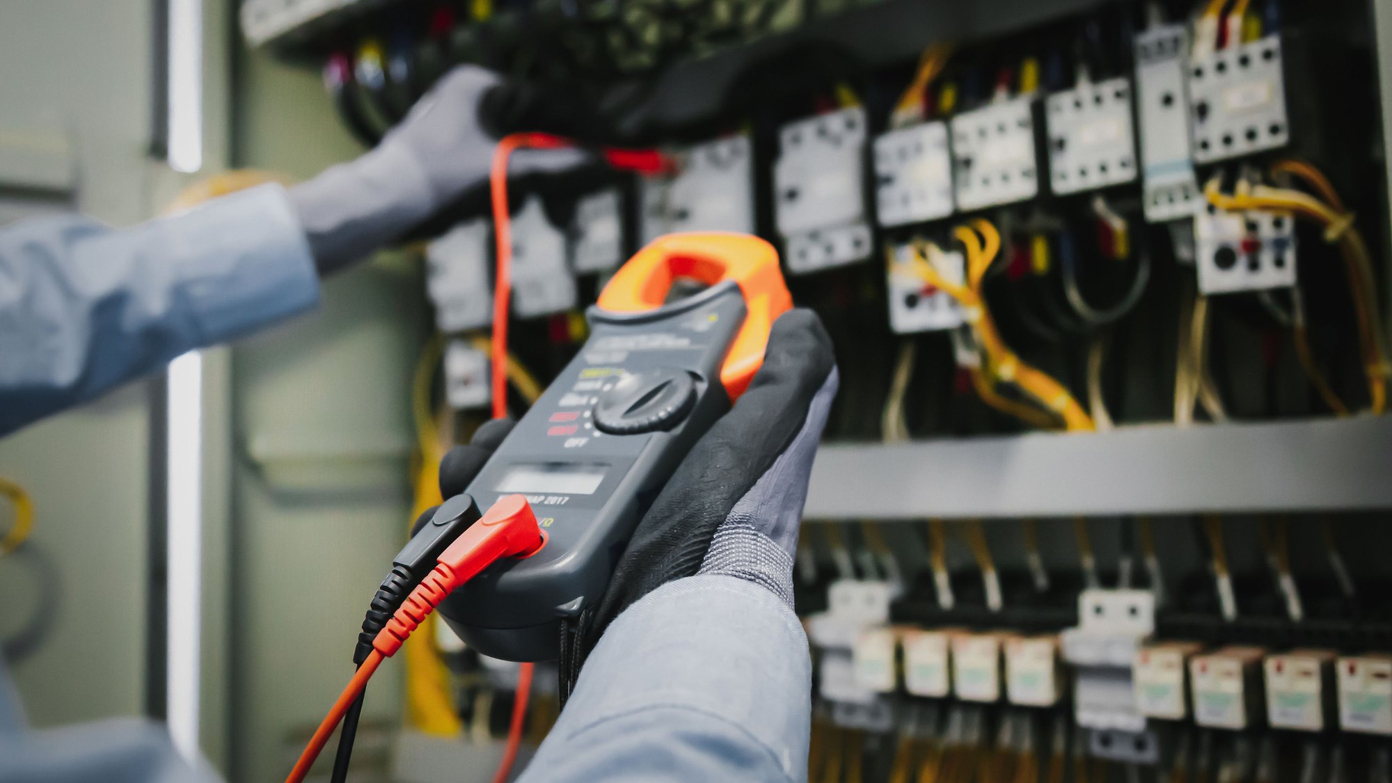 A gloved hand holding a multimeter probe against a row of circuit breakers inside an electrical panel