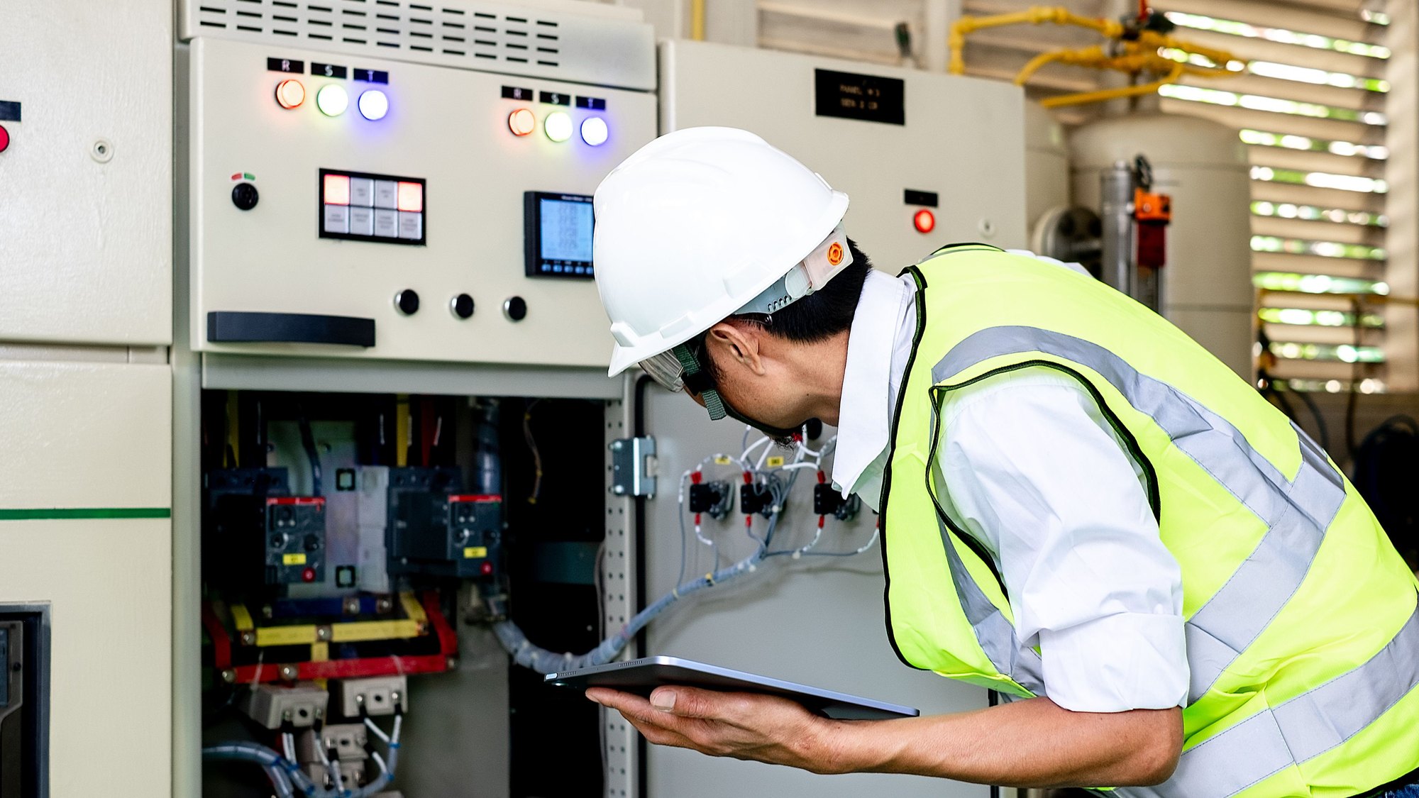 A technician in a hard hat and high-visibility vest inspecting switchgear with a tablet in hand