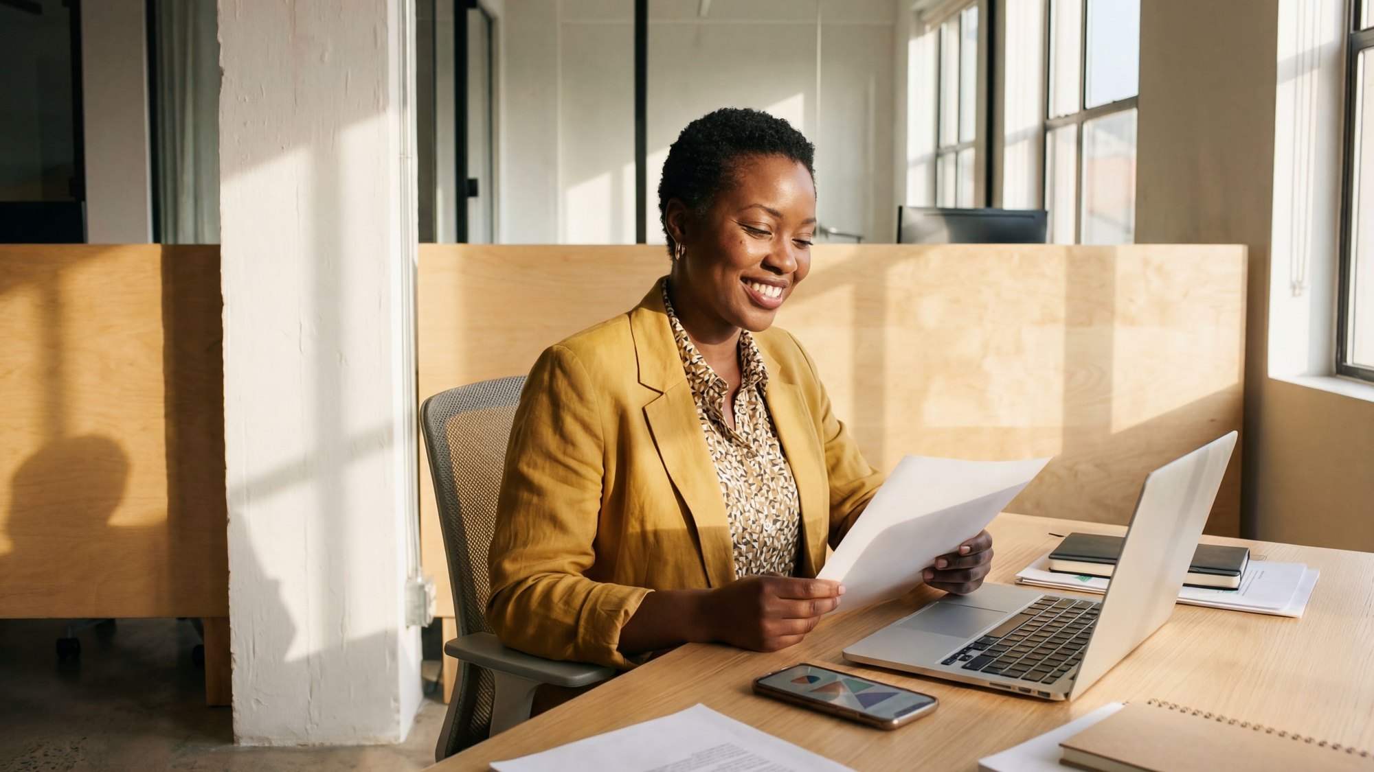 A woman in a blazer reading a printed report at her laptop in a bright office