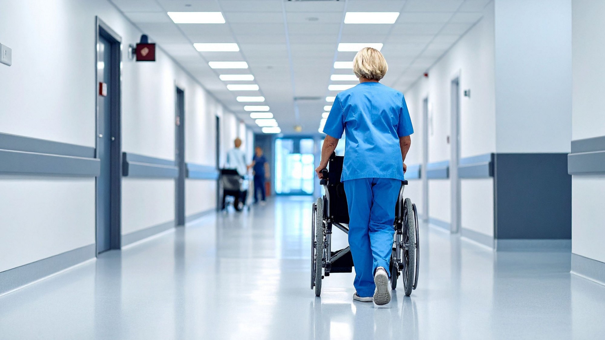 A nurse pushing a wheelchair away down a long hospital corridor