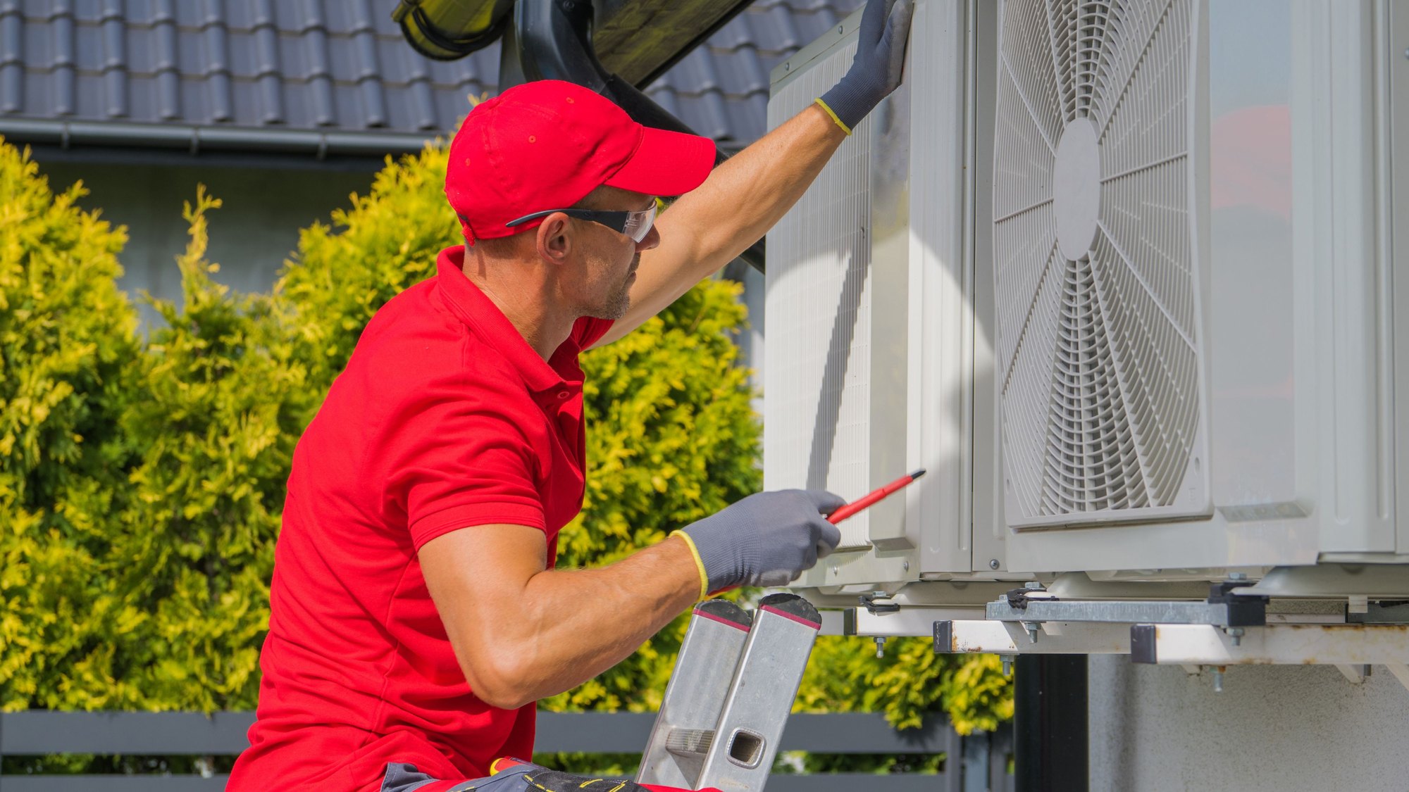 A technician in a red cap servicing an outdoor air conditioning condenser unit
