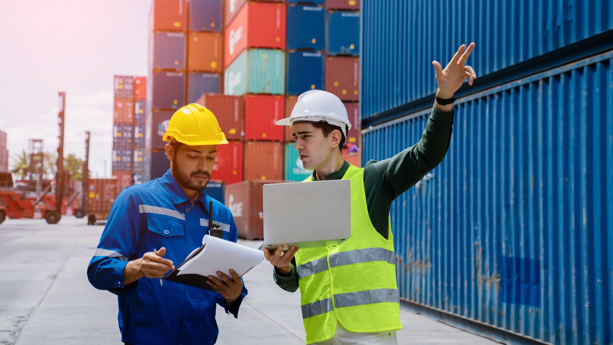 Two workers with a laptop and clipboard beside stacked shipping containers at a port