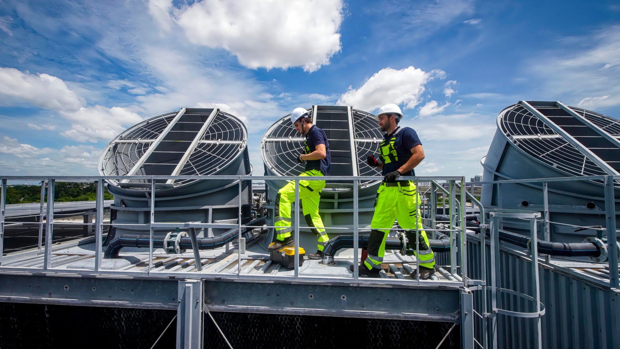 Two workers in high-visibility clothing on a rooftop walkway among large industrial fan units