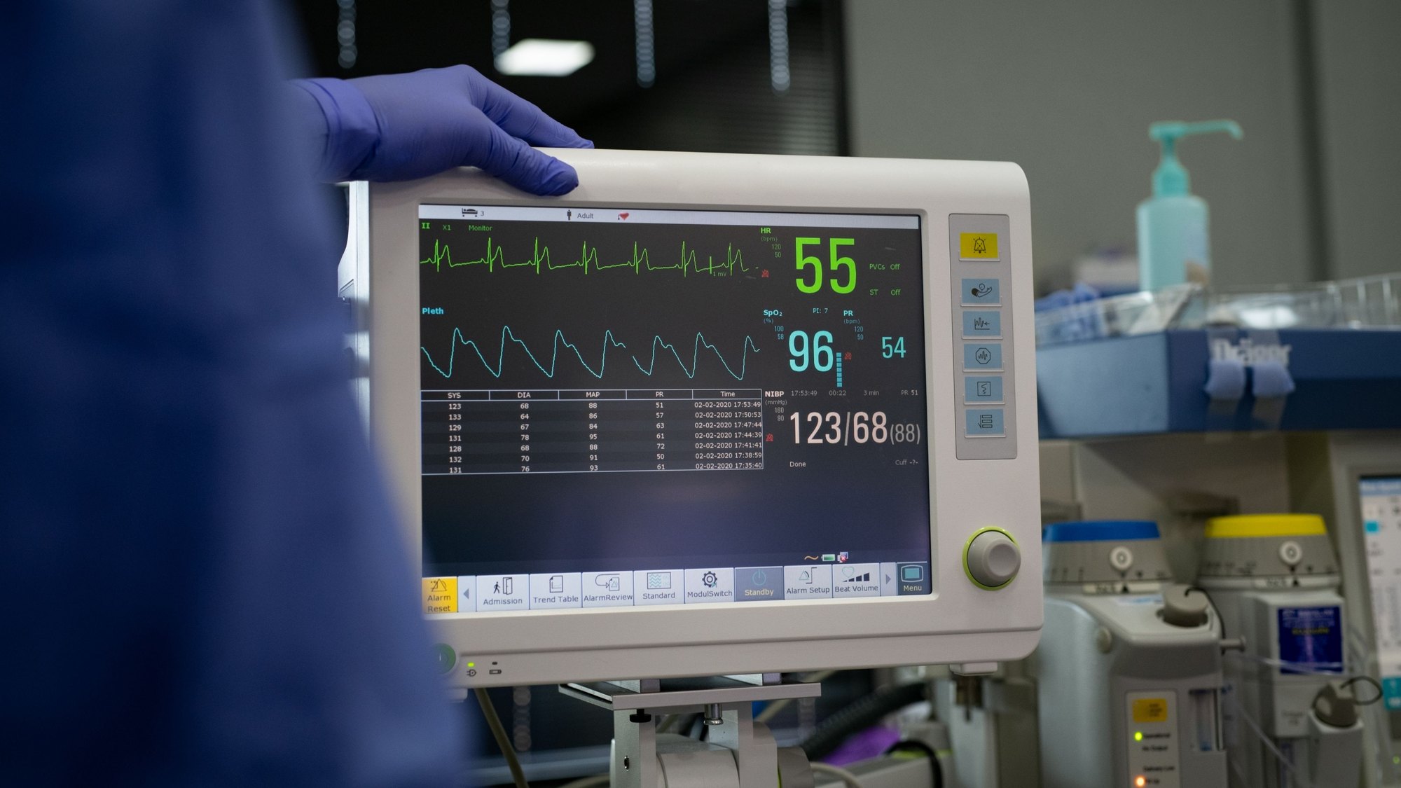 A gloved hand adjusting a bedside patient monitor showing heart rate and blood pressure