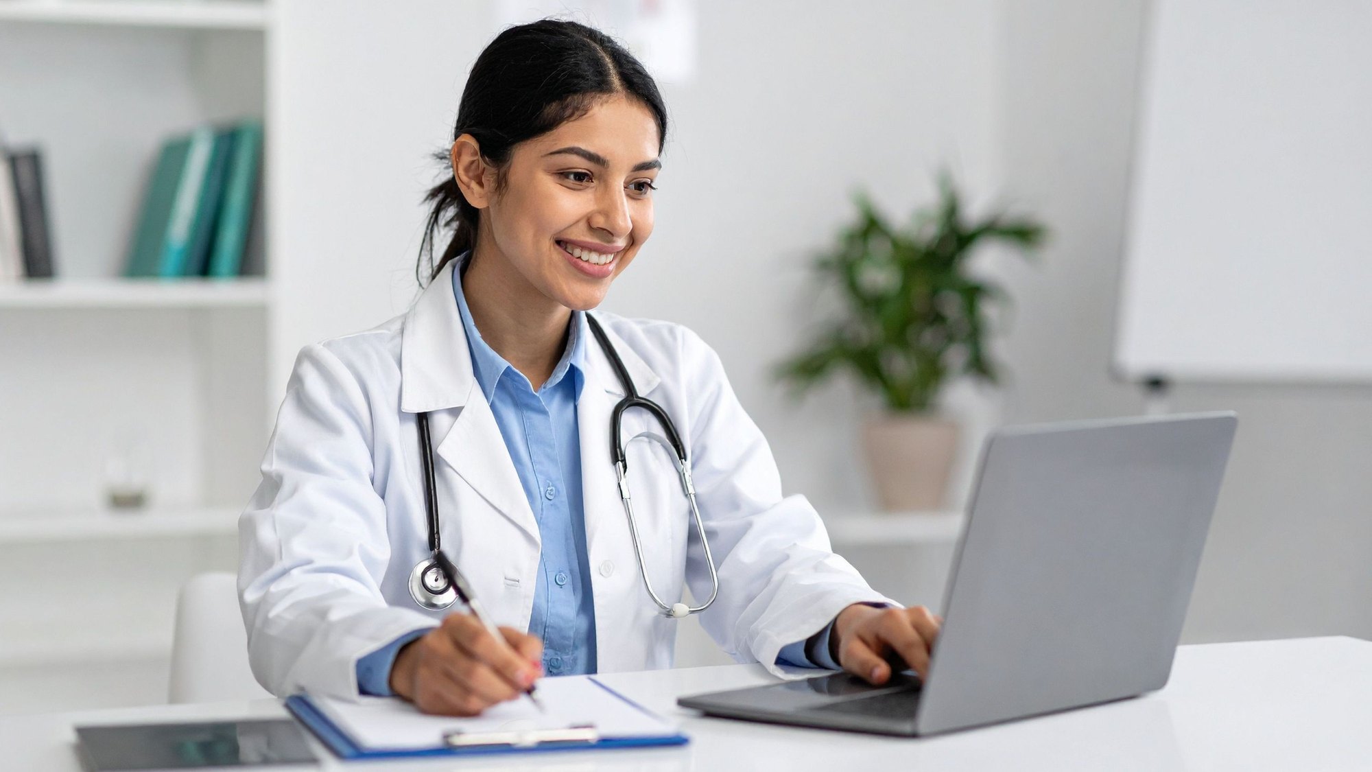 A clinician in a white coat and stethoscope taking notes at a laptop