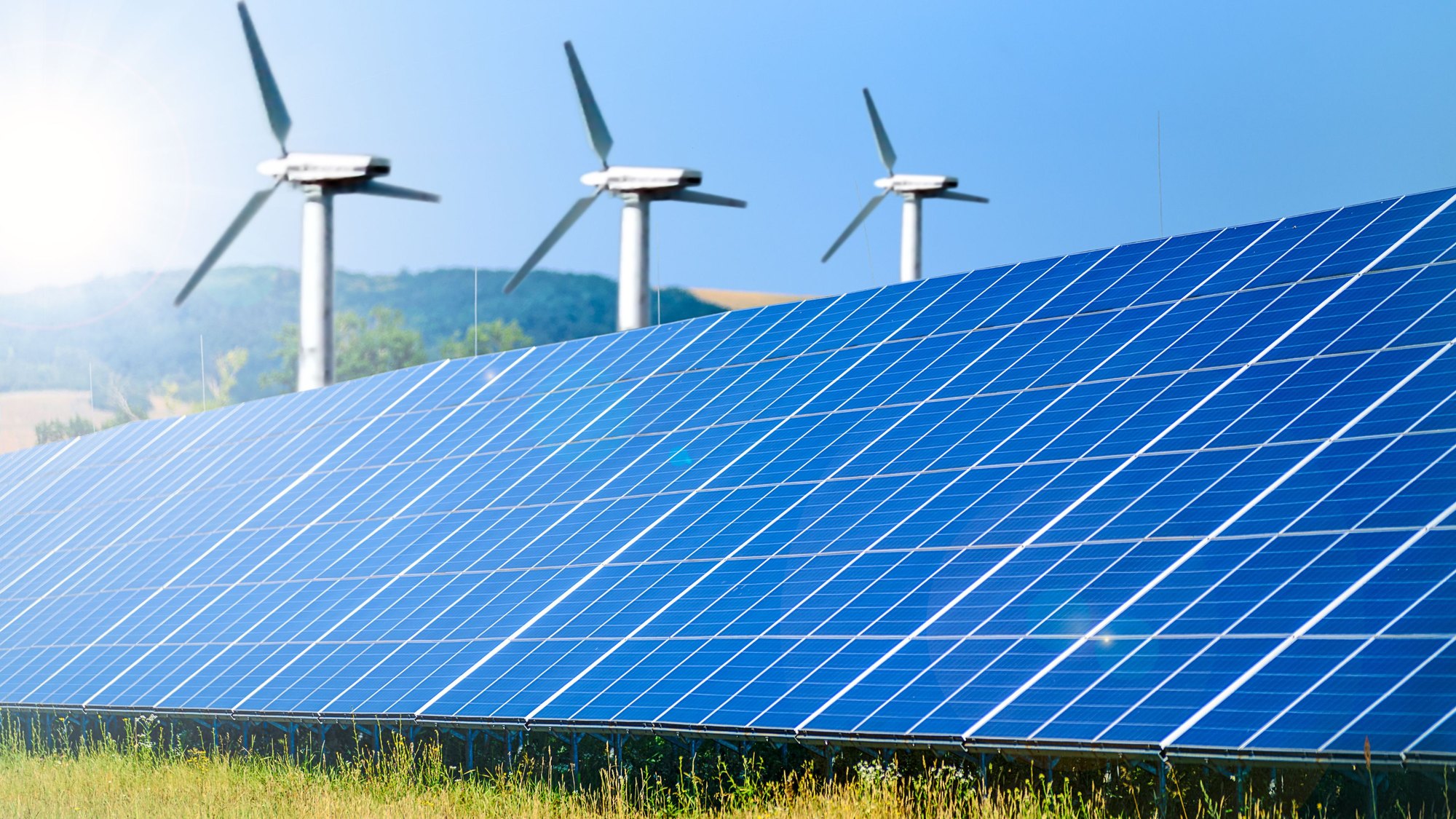 A solar array in the foreground with wind turbines on the hills behind it