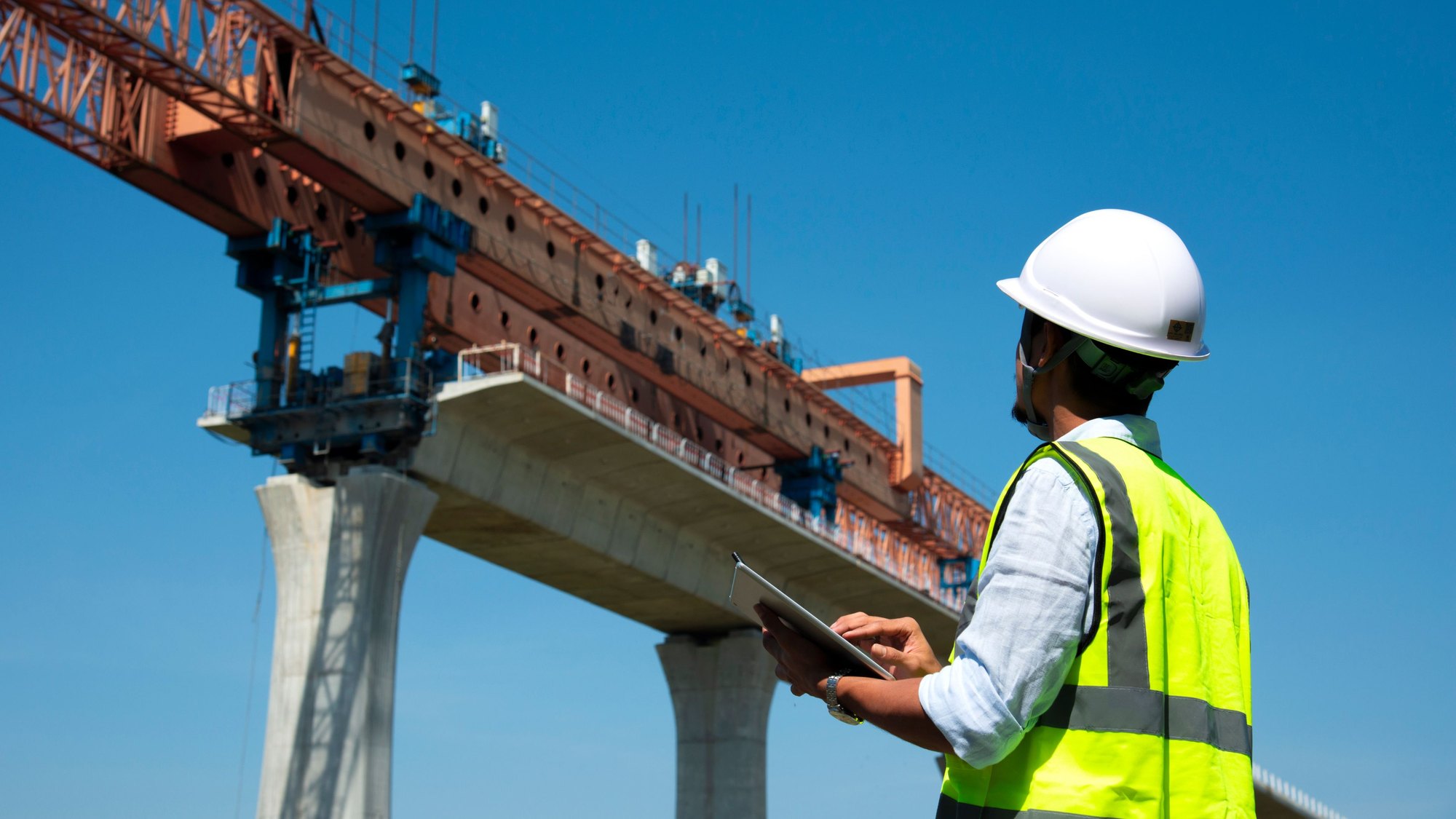 An engineer in a hard hat and high-visibility vest with a tablet, standing below a concrete bridge span under construction