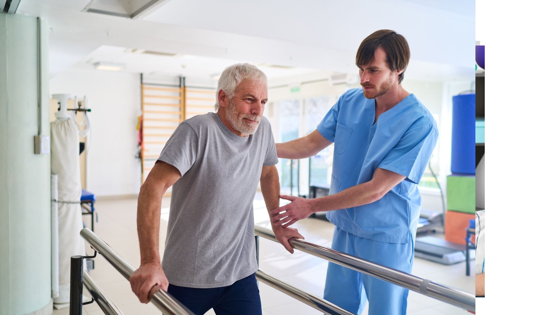 A physiotherapist in blue scrubs supporting an older man walking between parallel bars