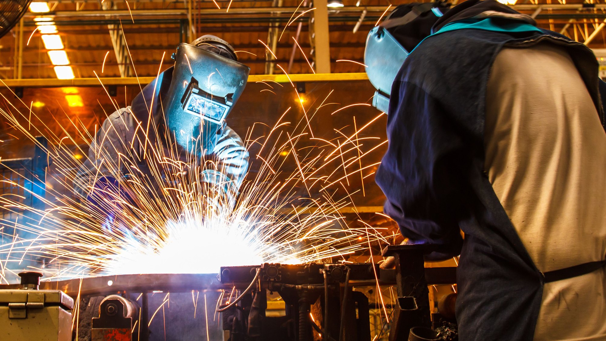 A welder in a helmet striking an arc, sparks flying across the workbench