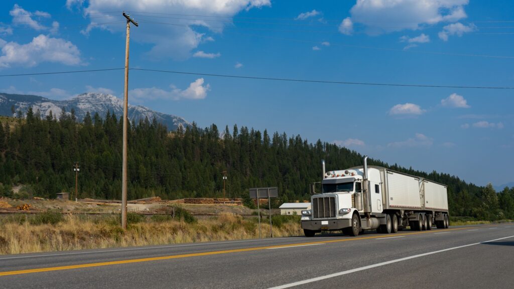 A white semi-truck on an open highway with forest and mountains behind it